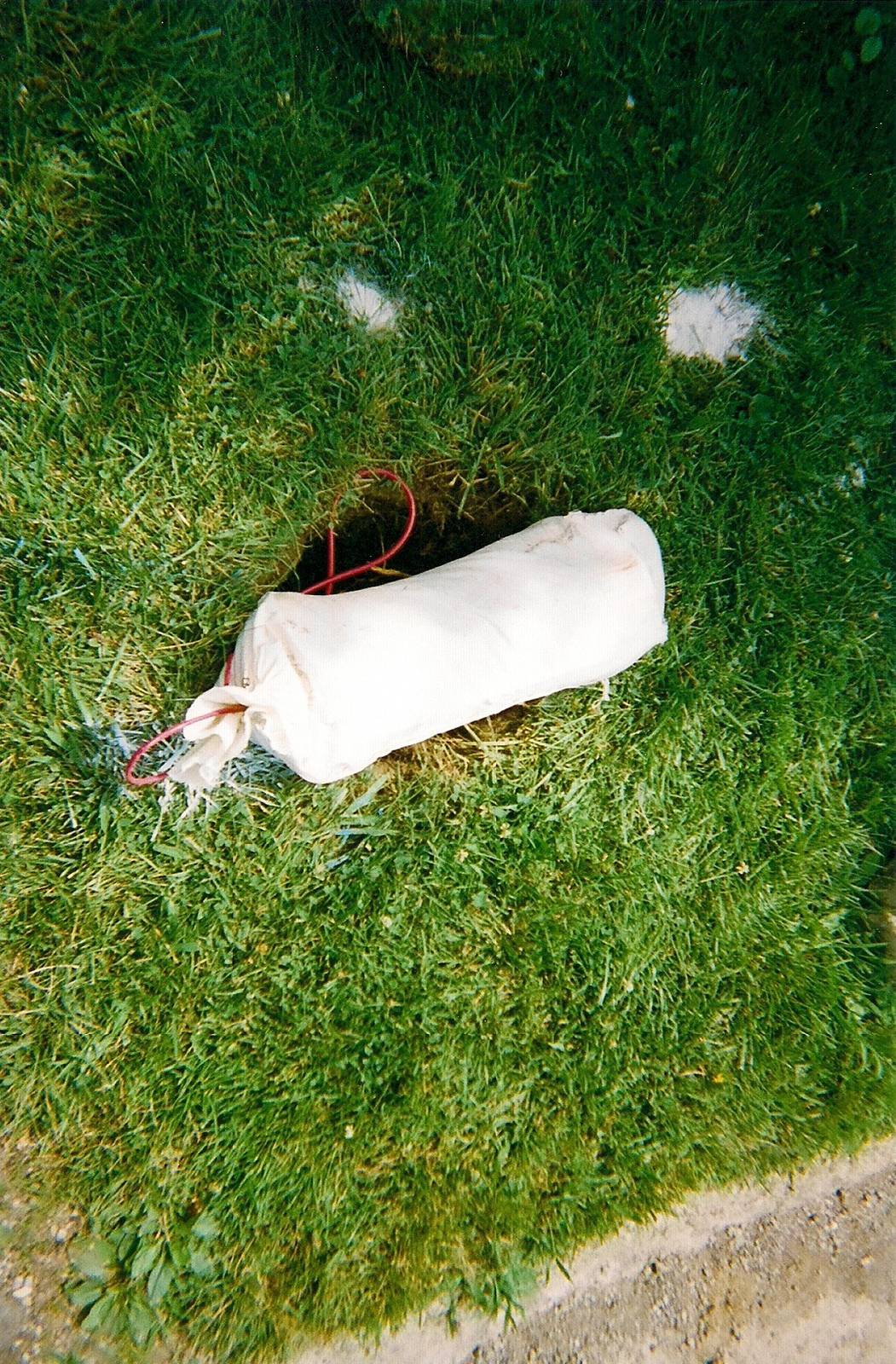 Packaged sacrificial anode with a red lead, on grass beside a hole ready for burial.