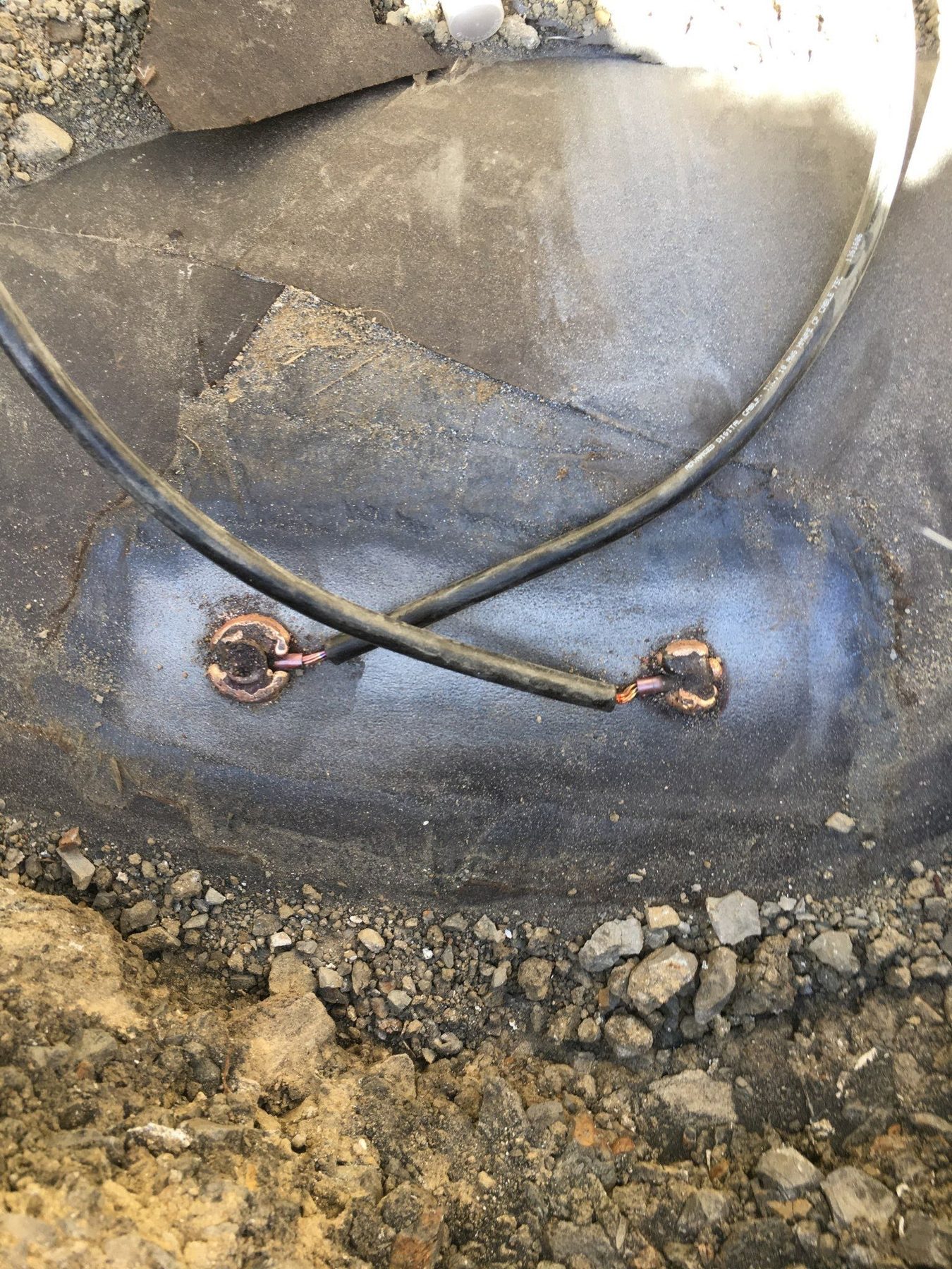 Black cables welded to a coated pipe in an excavation.