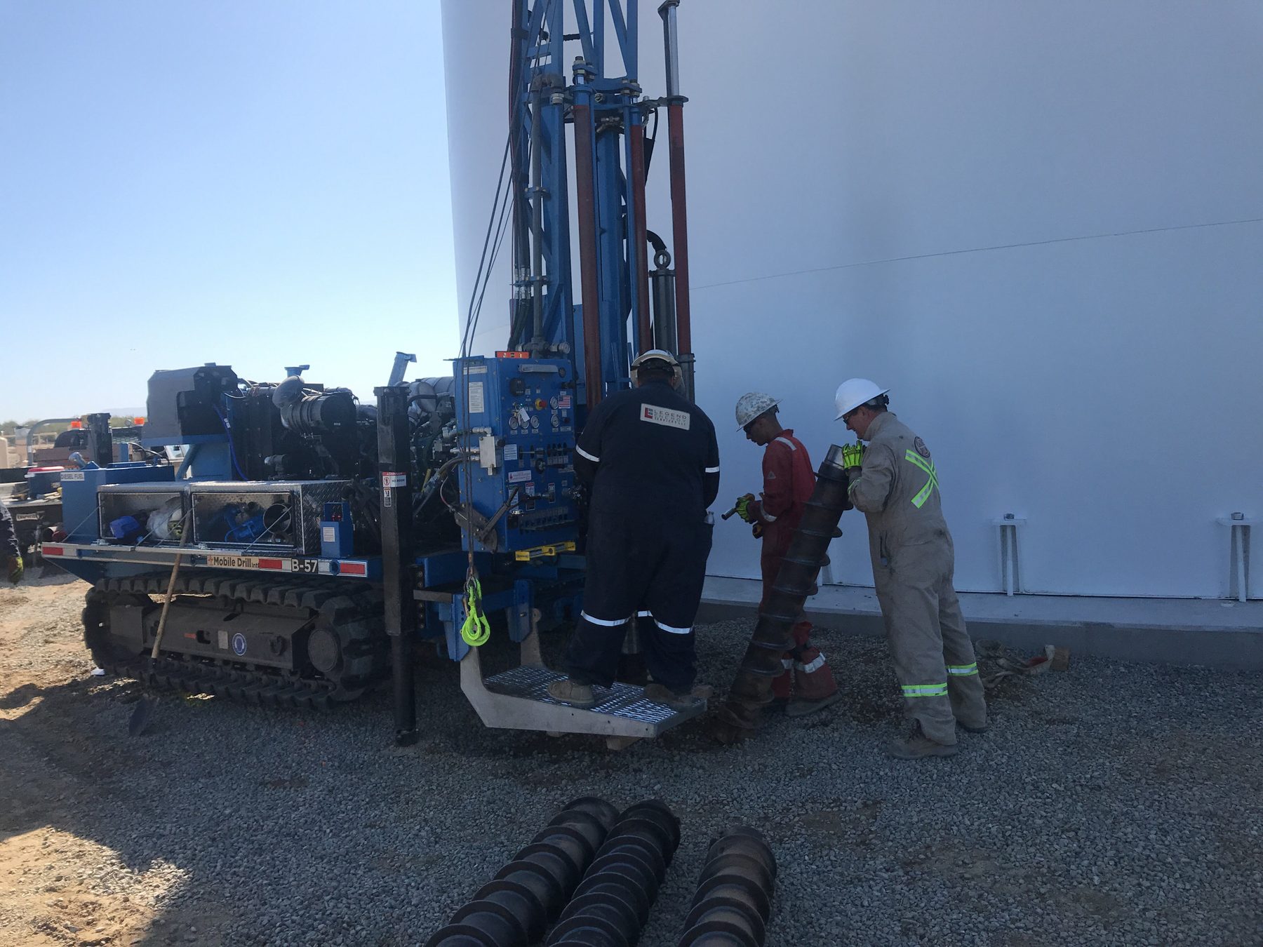 Track-mounted drill rig and crew beside a storage tank