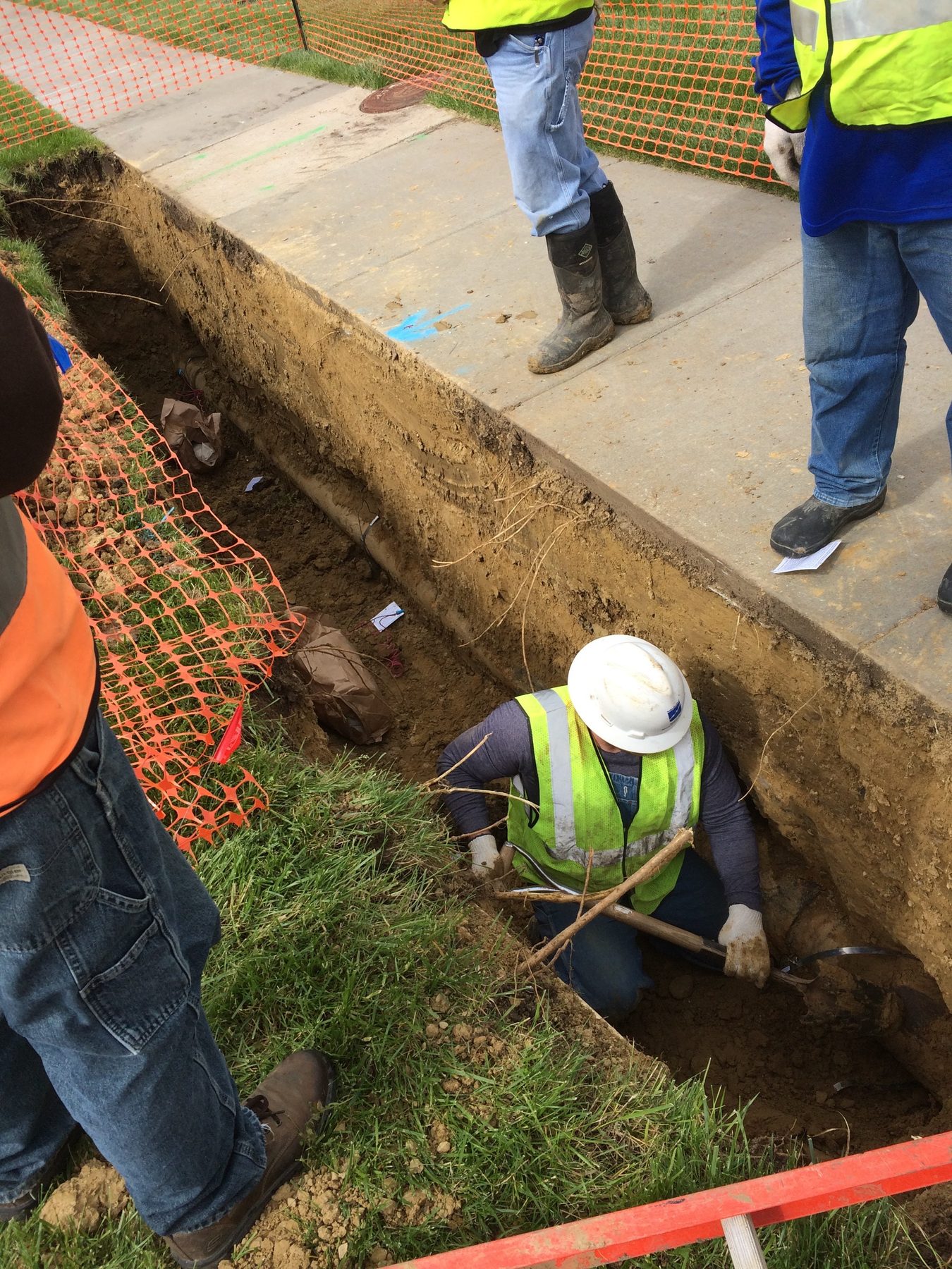 Worker in a white hard hat and hi-vis vest kneeling in a trench beside a sidewalk next to the exposed pipe, with orange safety fence and crew members standing above