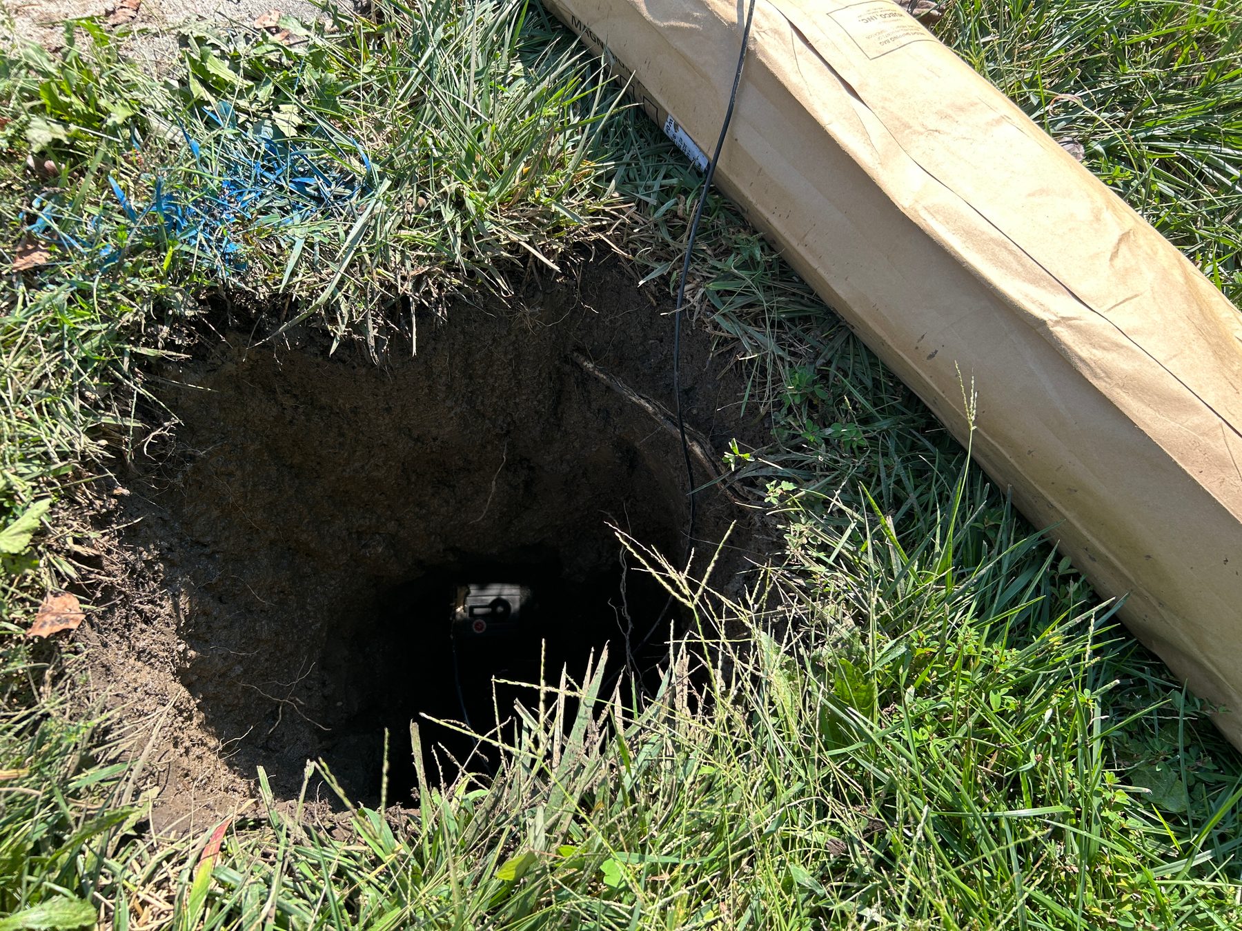 Small round keyhole excavation in grass with a lead wire running down to the cadweld attachment visible at the bottom, and a packaged anode lying alongside