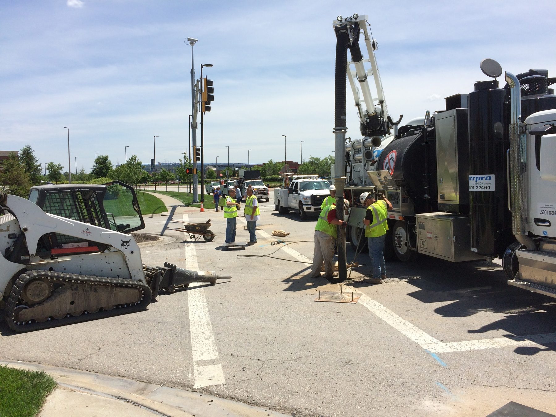 Hydrovac truck with its suction boom lowered into the pavement at a signalized intersection, crew in hi-vis vests working beside it and a skid steer with a hydraulic breaker in the foreground