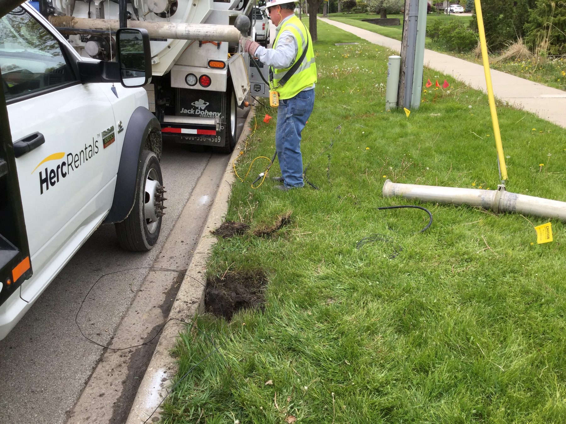 Worker in a hard hat and hi-vis vest at the back of a hydrovac truck parked along a curb, with small excavations cut into the grass at the curb edge, wire leads and a suction tube lying on the lawn