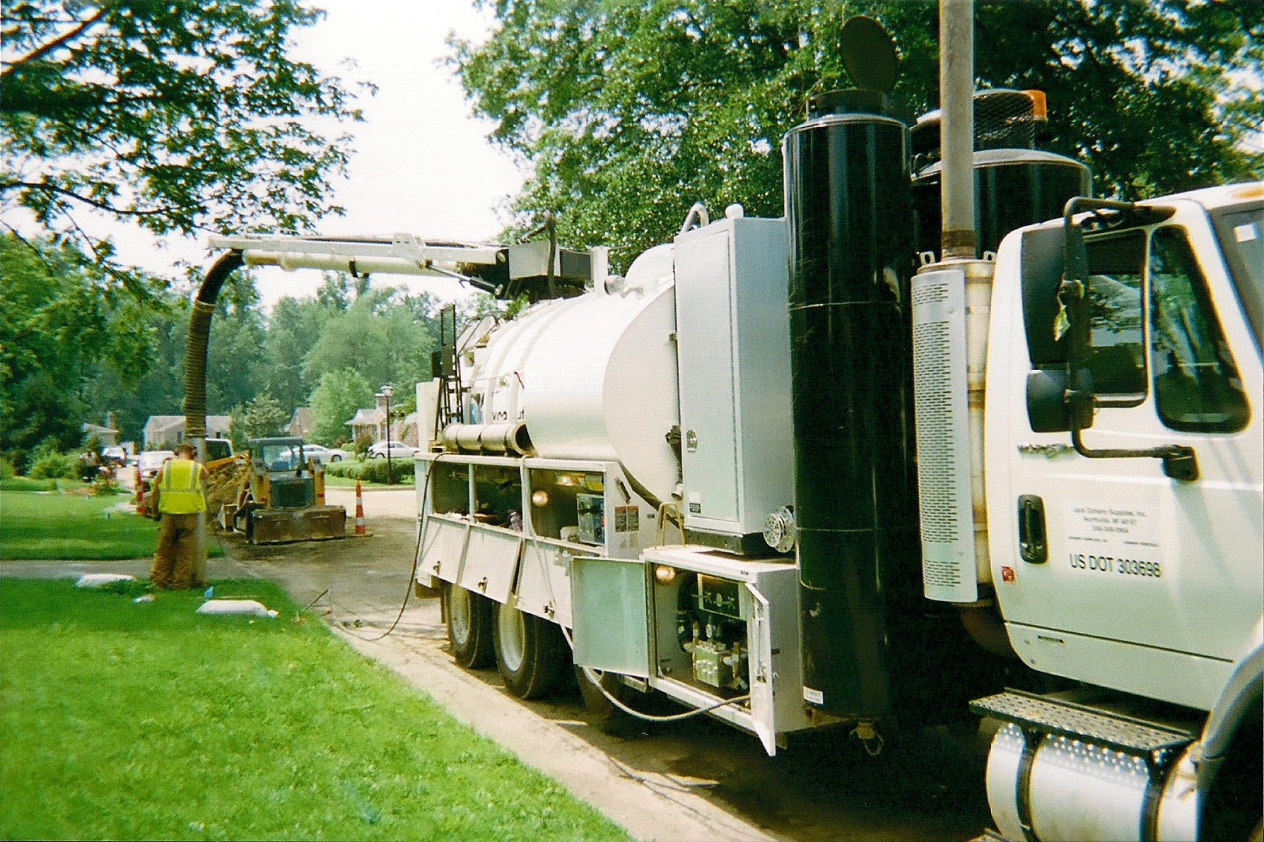 Hydrovac excavation truck parked along a tree-lined residential street, its suction boom extended to an excavation where a worker in a hi-vis vest stands beside a skid steer