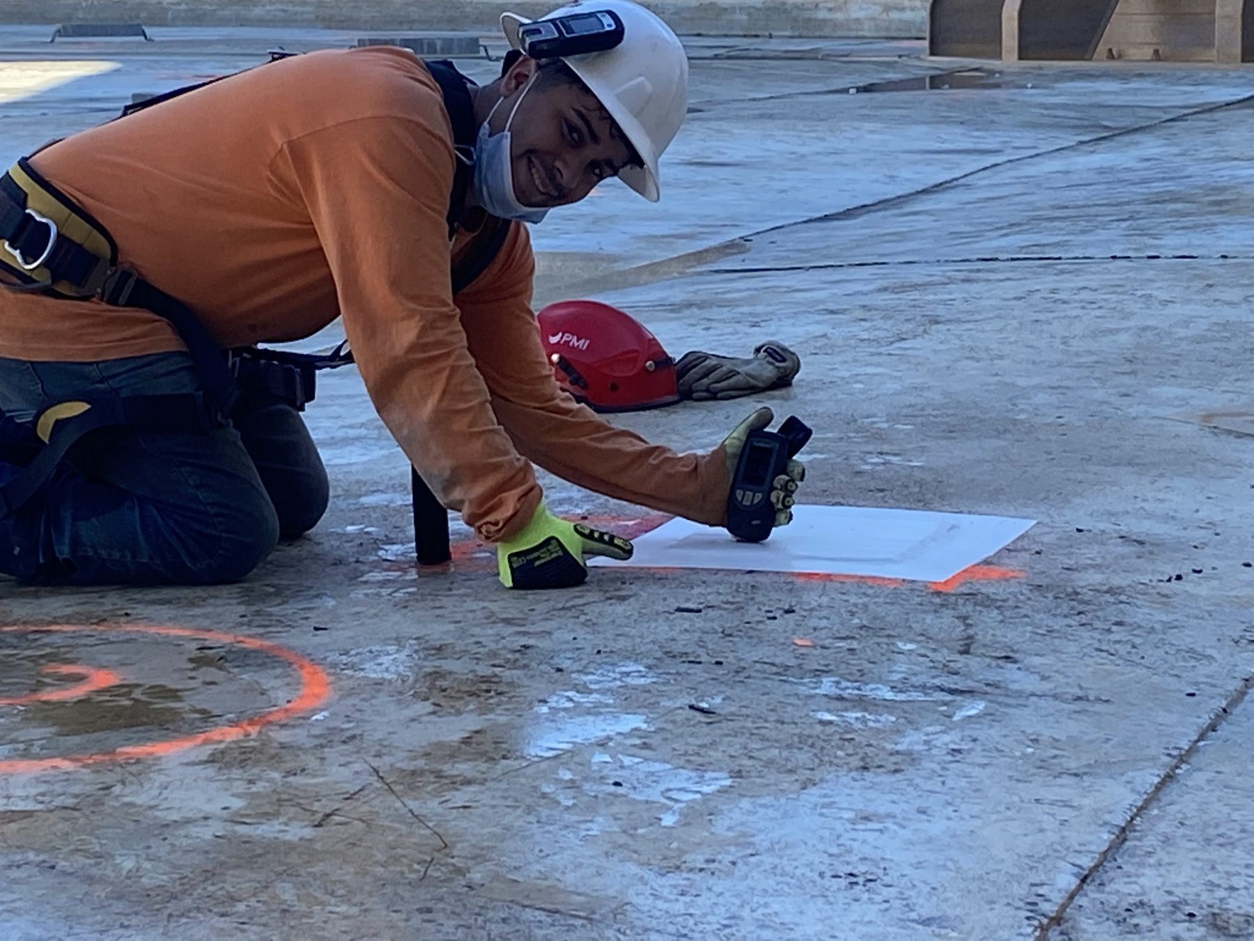 Worker in a hard hat and safety harness kneeling on concrete with a handheld survey instrument.