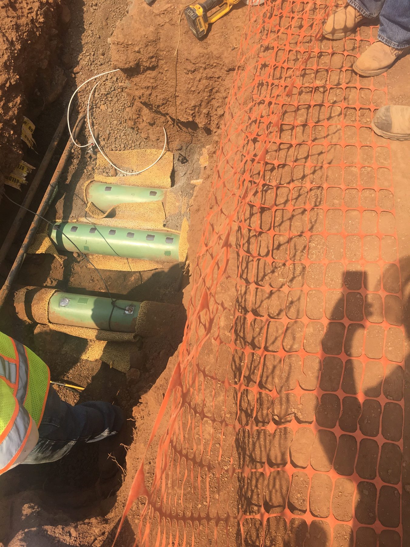 Green pipes in an open trench with yellow padding and orange safety fence.